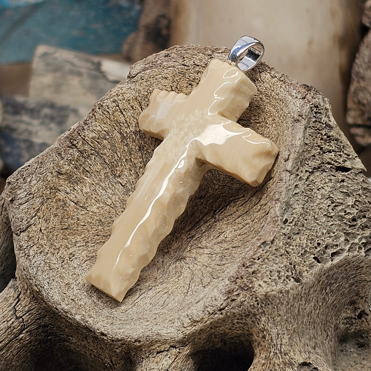 Woolly Mammoth fossil ivory Christian cross pendant resting on bone, showing light mineralization and a silver bale, crafted by Kristin R Park at Ice Age Treasures-Boneyard Alaska Materials