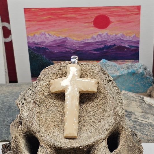 Woolly Mammoth fossil ivory Christian cross pendant displayed on a mammoth tusk with a mountain painting in the background, crafted by Kristin R Park at Ice Age Treasures.